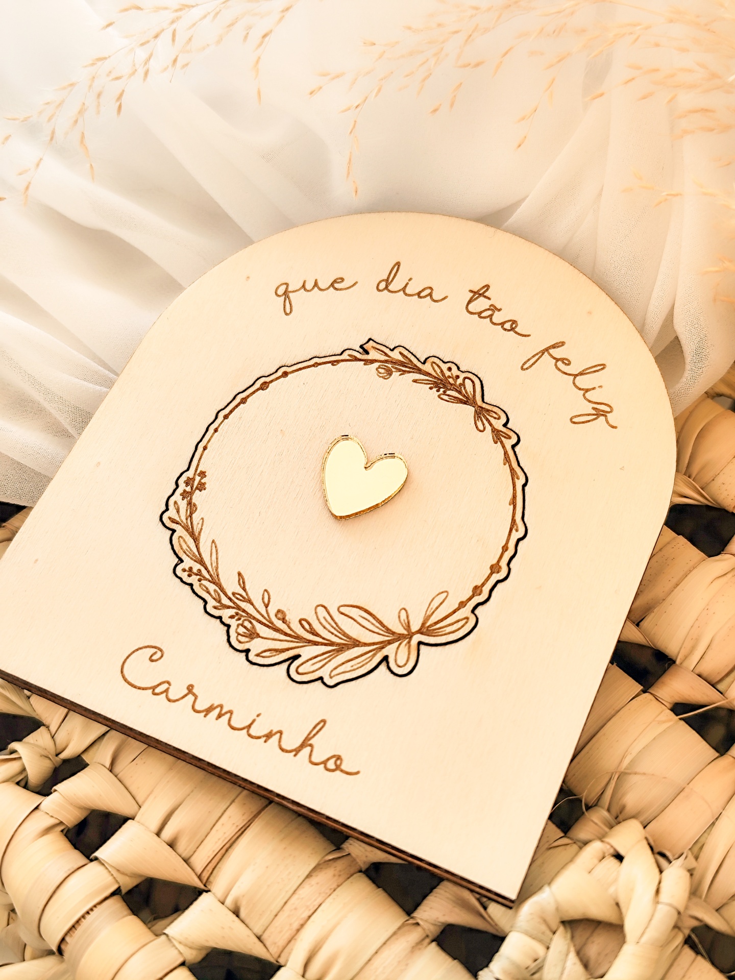Placa decorativa de madeira com coração dourado e texto gravado, sobre fundo de tecido branco e cesta de palha