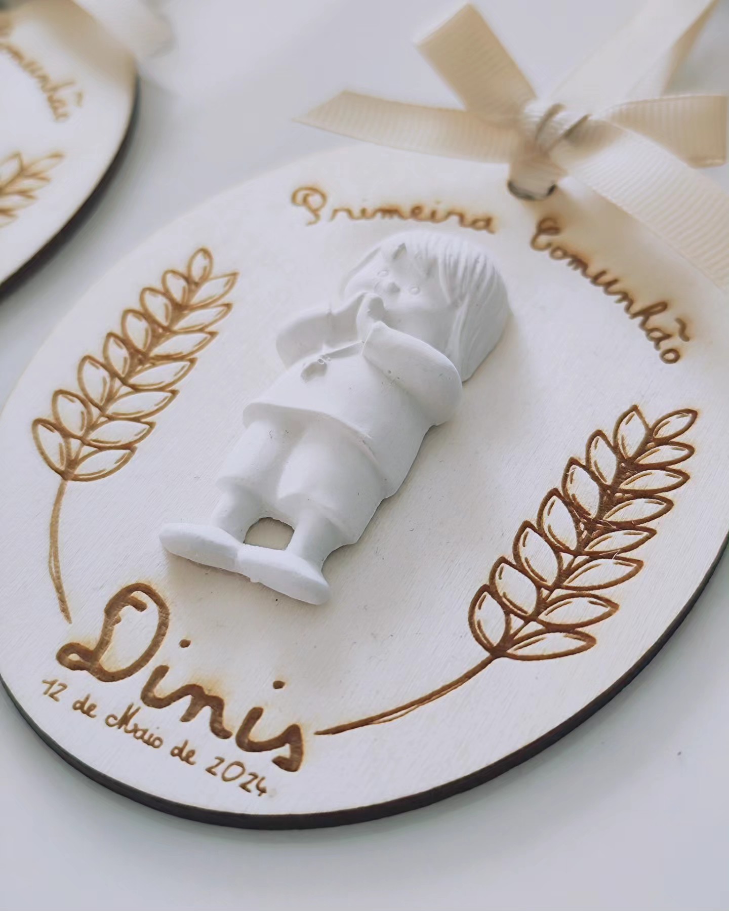 Plaqueta oval branca com figura de menino e texto dourado sobre primeira comunhão