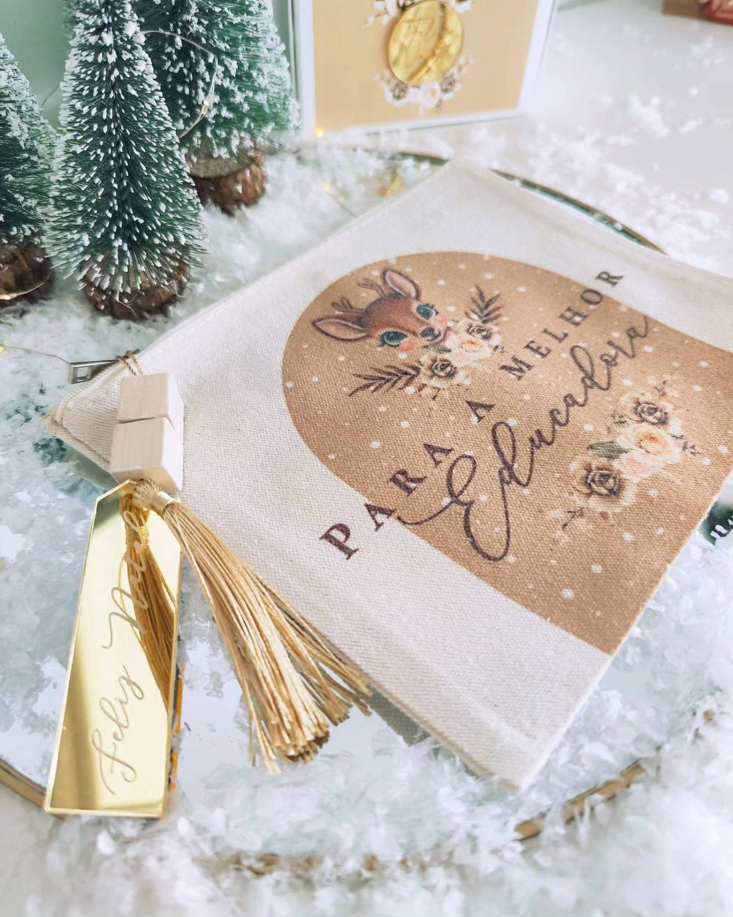 Saco tecido bege com estampa de veado e flores e texto para melhor educadora, etiqueta dourada félicitando Natal, mini árvores Natal e neve artificial
