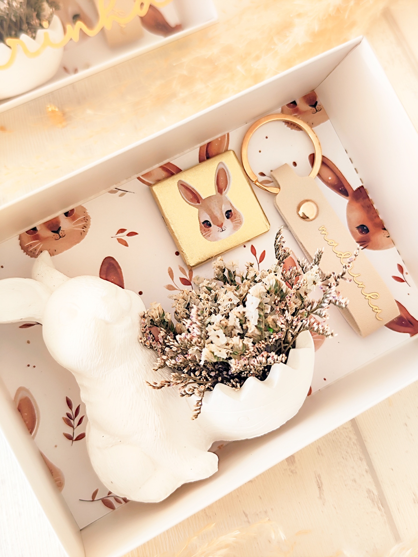 Conjunto com figura de coelho cerâmico, chaveiro de couro bege e cubo decorativo de coelho dentro de caixa branca com ilustrações.