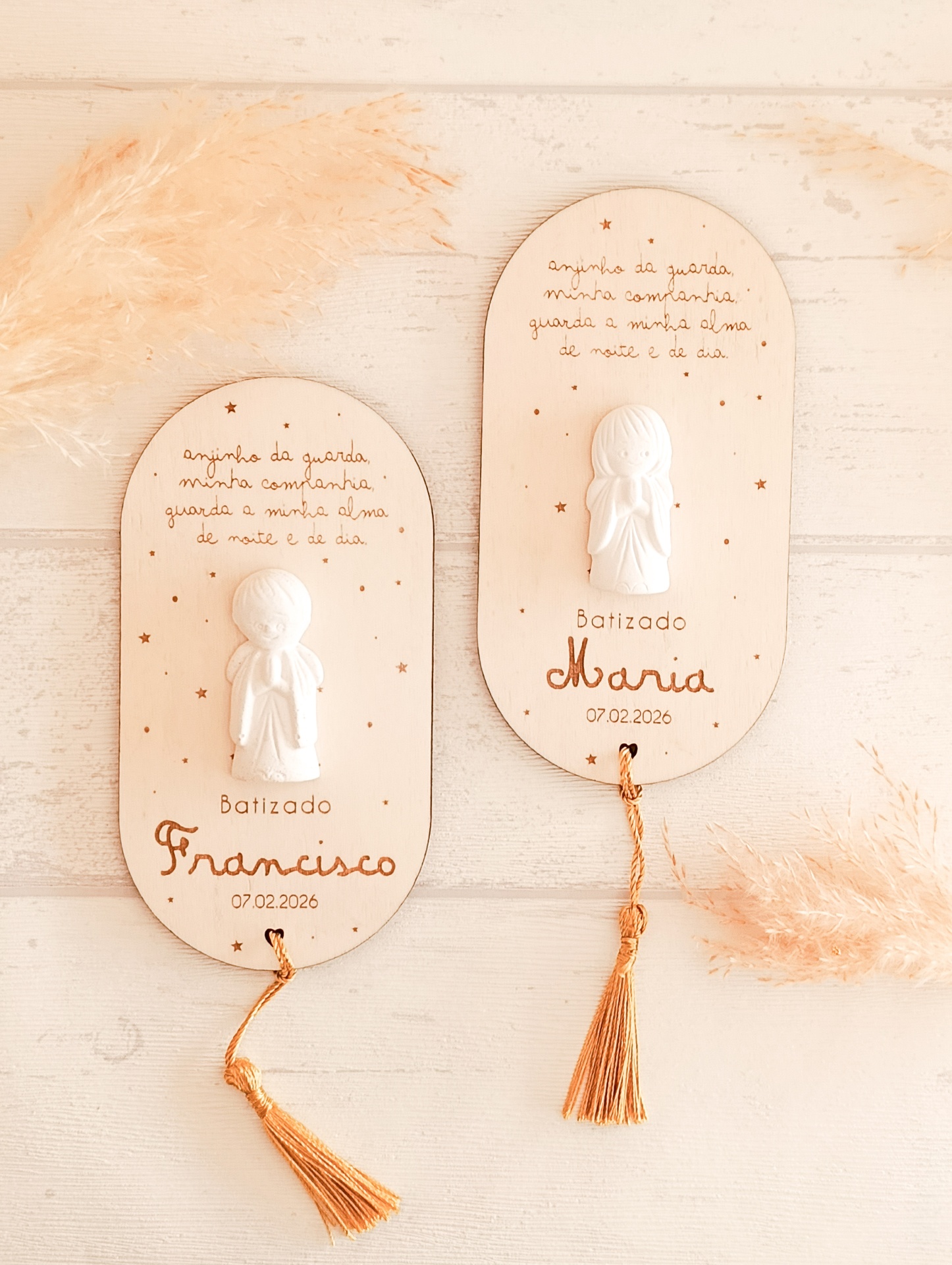 Dois quadros decorativos de madeira clara com anjos em relevo branco e franjas douradas