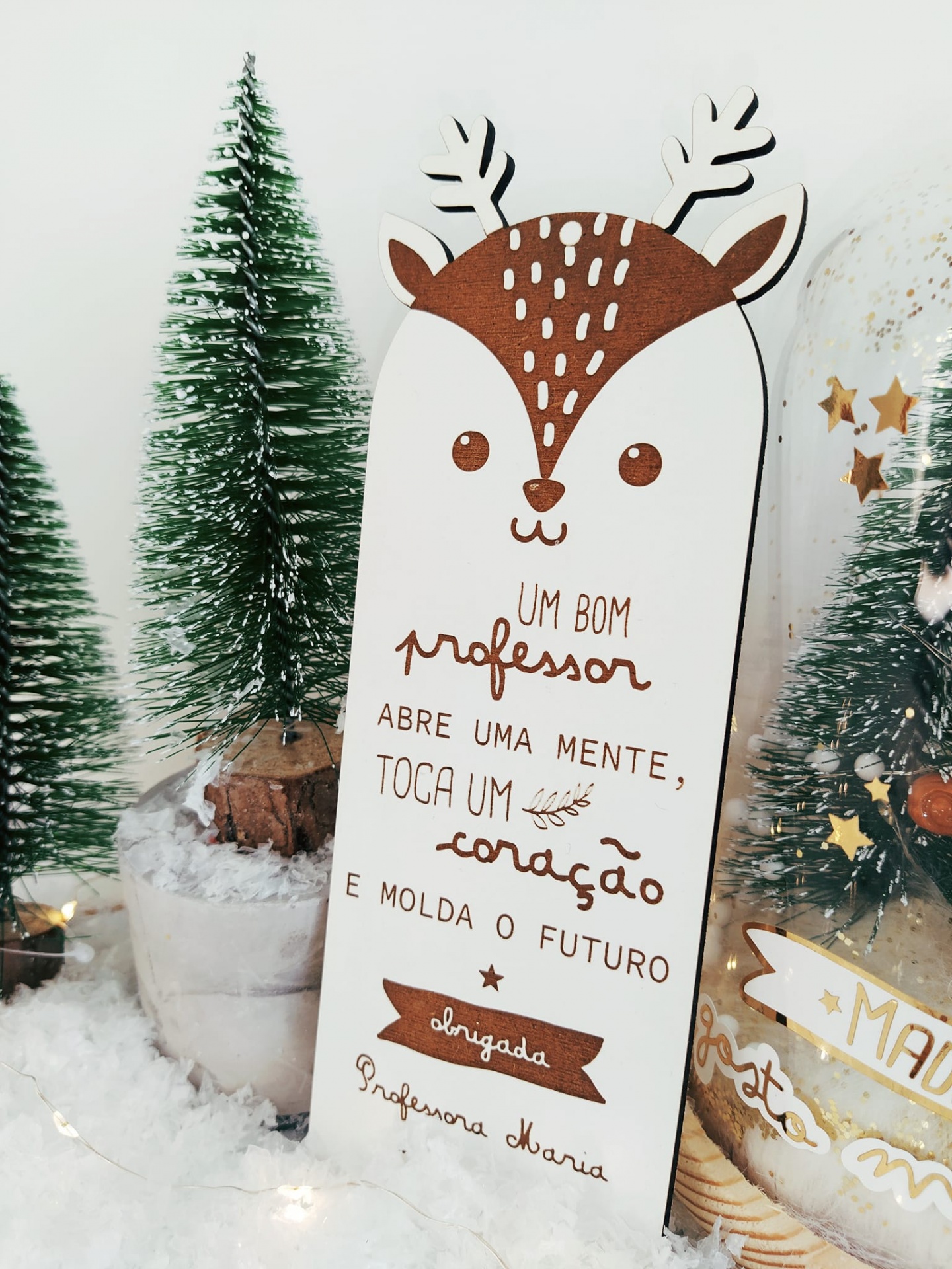 Placa decorativa de rena com texto para professora entre árvores e neve artificial
