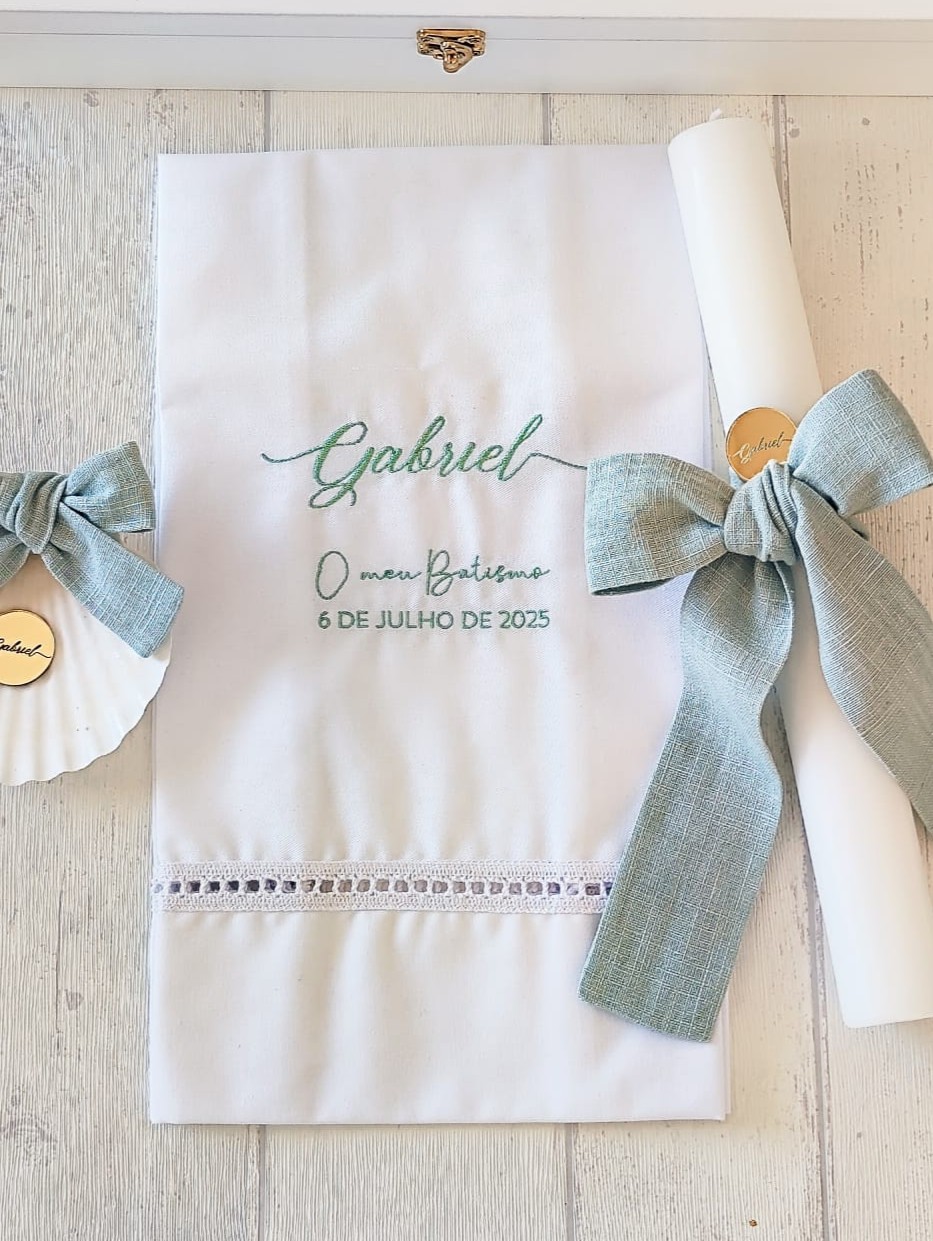 Toalha branca com bordado verde, rolo branco e concha decorados com laços verdes num fundo de madeira clara