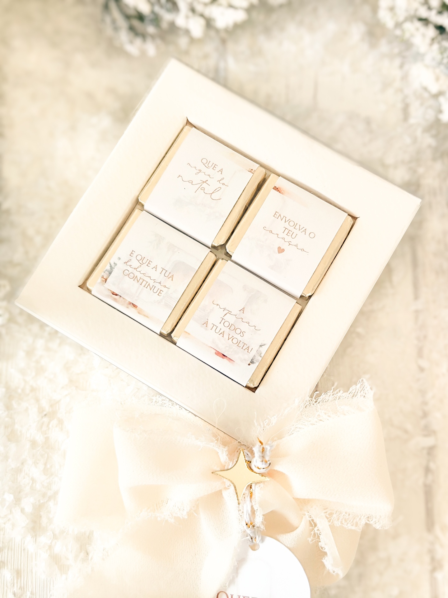 Caixa branca com quatro chocolates pequenos embrulhados em papel creme com frases em português e decorada com laço creme e estrela dourada.