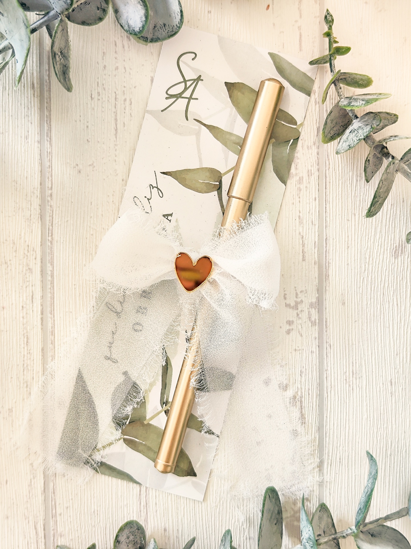 Caneta dourada com laço branco e cartão decorativo com folhas verdes
