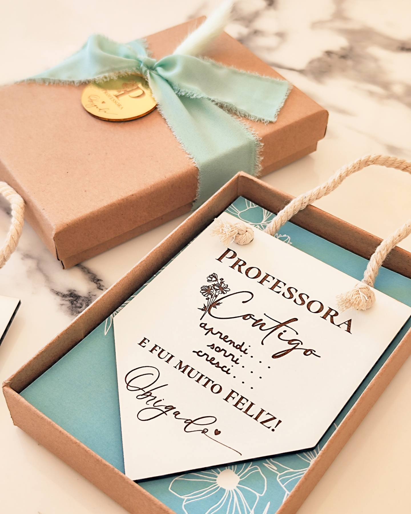Placa decorativa branca numa caixa castanha com fita verde-água, cartão azul floral e selo dourado
