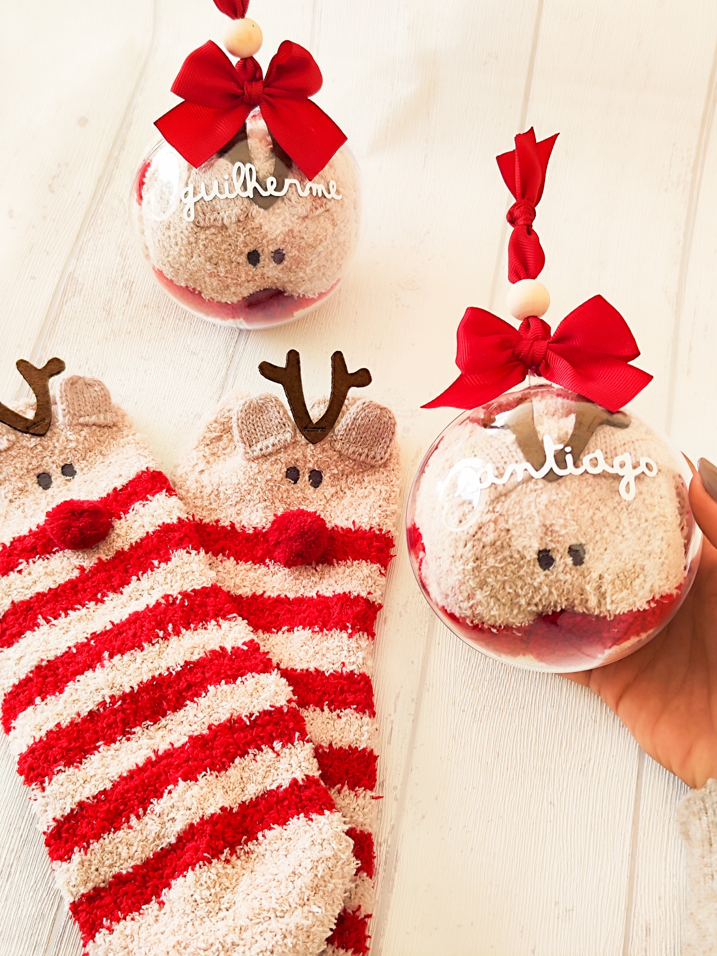 Meias peludas com padrão de riscas vermelhas e creme e decoração de rena dentro de bolinhas transparentes com nomes.