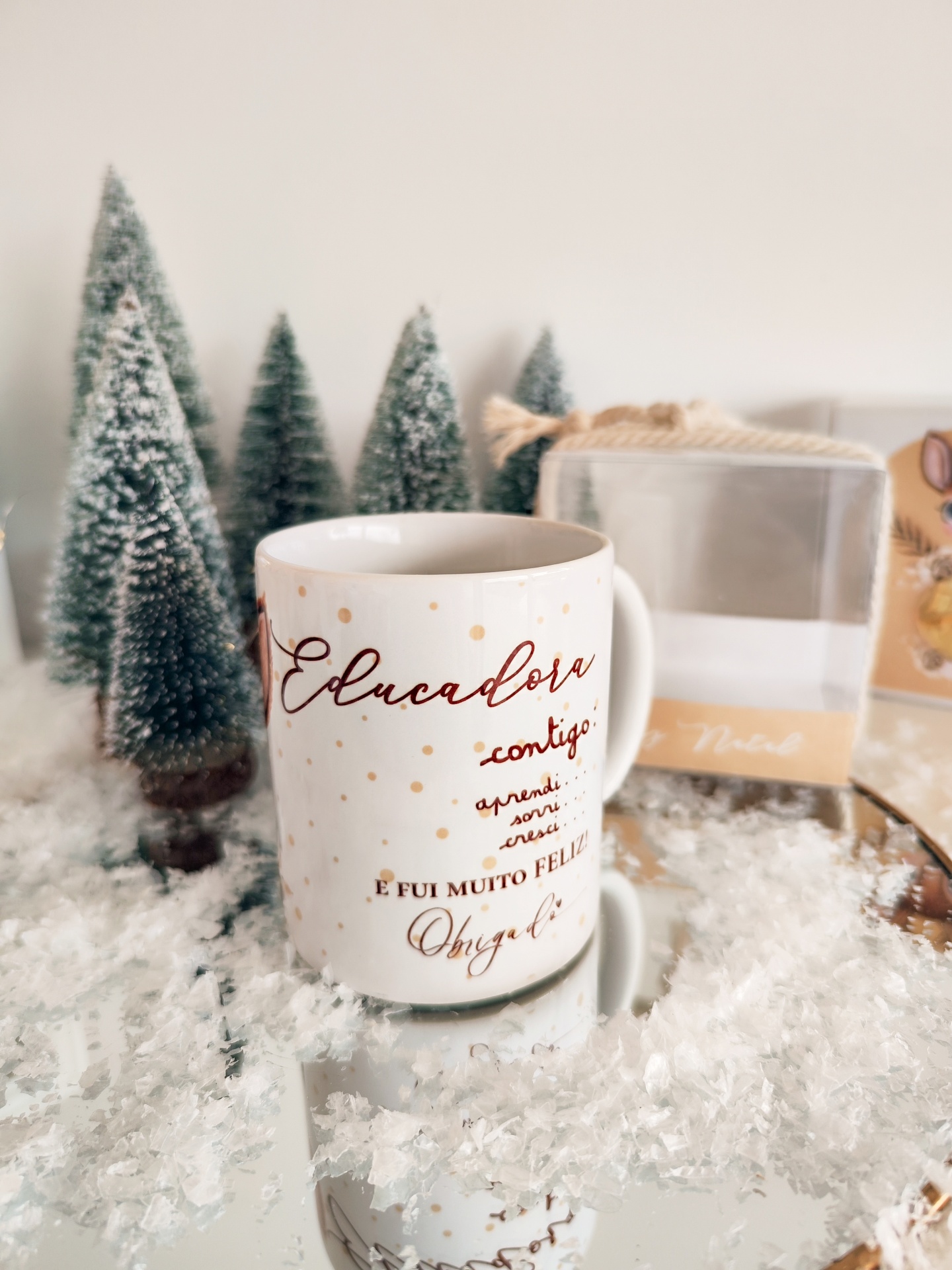 Caneca branca decorativa com texto em castanho, acompanhada de miniaturas de árvores de Natal e neve falsa