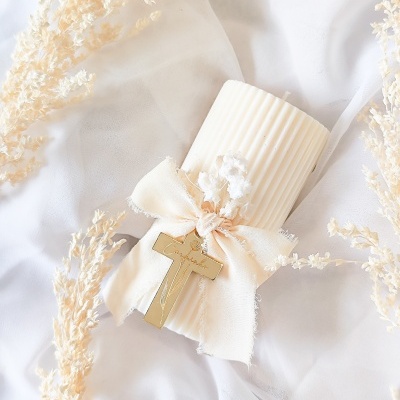 Vela branca canelada com decoração de laço, cruz dourada e flores secas sobre tecido branco.