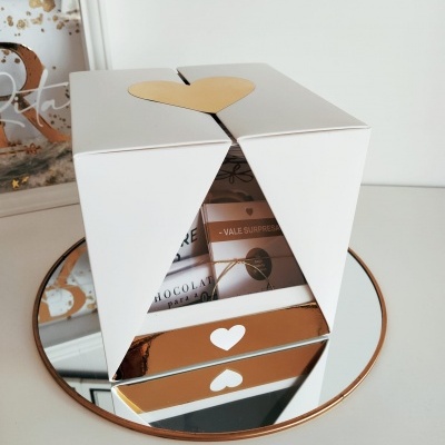 Caixa branca com coração dourado, sobre espelho redondo dourado, com vale-surpresa e embalagem de chocolate visíveis.