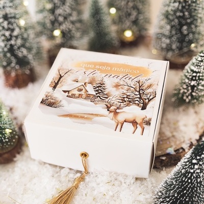 Caixa branca decorativa com ilustração natalícia e pompom dourado em ambiente com árvores de Natal