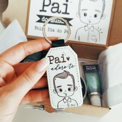 Chaveiro branco com ilustração e texto 'Pai adoro-te' segurado por mão feminina frente a caixa de presente.