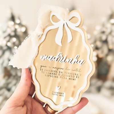 Placa decorativa dourada com laço branco e texto em relevo sobre tema Natal