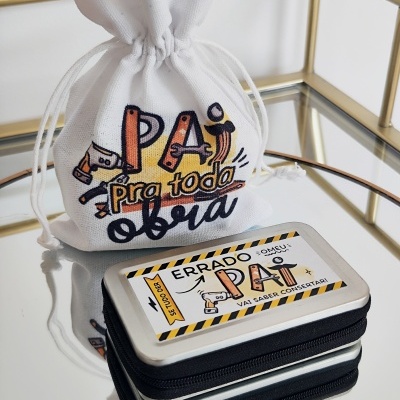 Saco branco estampado com mensagem para pai e caixa metalizada com texto humorístico, sobre superfície espelhada