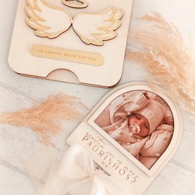 Convite de madeira claro com asas douradas e foto de bebé com laço branco