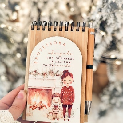Bloco de notas em madeira com desenho de lareira e personagens, texto dedicado a professora, caneta de madeira ao lado.