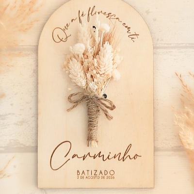Placa decorativa em madeira clara com ramo de flores secas e texto gravado para batizado.