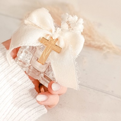 Frasco de vidro com laço bege e cruz dourada nas mãos femininas