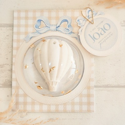 Produto decorativo balão branco com detalhes dourados em cartão xadrez bege e branco com etiqueta azul e branca.