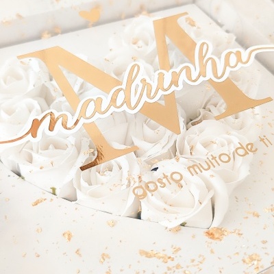 Caixa transparente com flores brancas artificiais, letra 'M' e texto dourado 'madrinha' e 'gosto muito de ti'