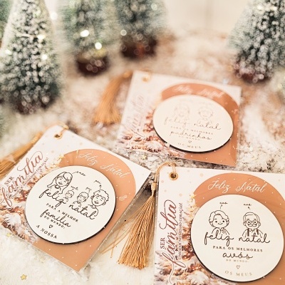 Enfeites de Natal brancos com ilustrações de família e texto em cartões castanhos com franjas douradas sobre neve artificial com pinheiros