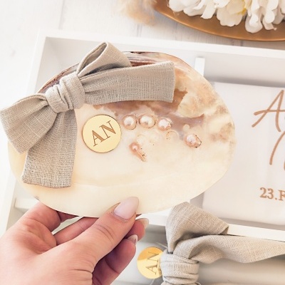 Concha decorativa com laço bege, pérolas e placa dourada com letras AN