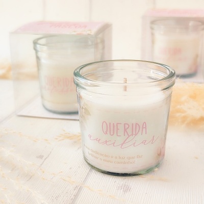 Vela em copo de vidro com texto rosa 'QUERIDA auxiliar' em cima de mesa de madeira branca