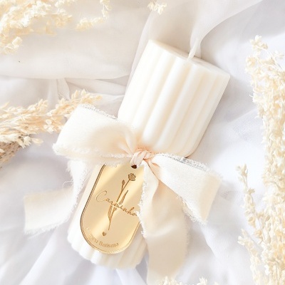 Vela branca canelada com fita creme e etiqueta dourada sobre tecido branco com flores secas