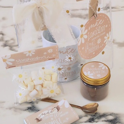 Conjunto de presente com saco transparente, marshmallows, frasco, caneca e barra de chocolate decorados com tema floral e tons rosa claro.
