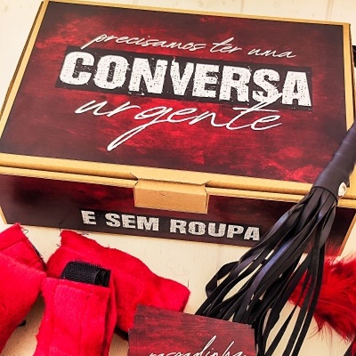 Caixa de cartão com texto, algemas vermelhas, instrumento de couro preto e livro 'raspadinha KAMASUTRA'