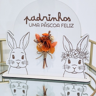Placa decorativa branca com desenhos de coelhos e texto de Páscoa com flores secas