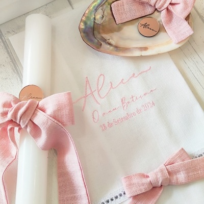 Toalha branca bordada com nome e data, vela branca e concha com laço rosa