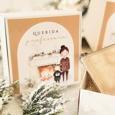 Caixa decorativa com ilustração e texto para professora junto a mala dourada e neve falsa