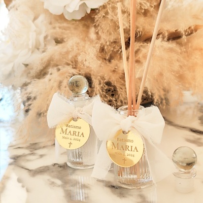 Dois frascos de vidro decorados para batismo com etiquetas douradas e laços brancos, sobre superfície de mármore e decoração floral em fundo. 