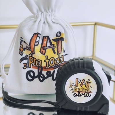 Fita métrica preta e saco branco com texto 'PAI pra toda a obra' em superfície espelhada e moldura dourada