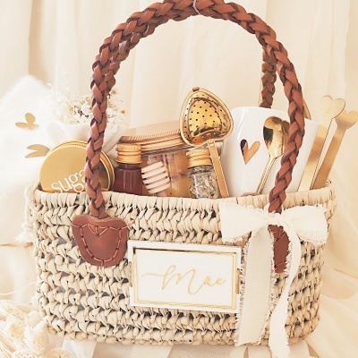 Cesto de palha com alças de couro e diversos itens para chá incluindo colher de chá em forma de coração e caneca branca com coração recortado.