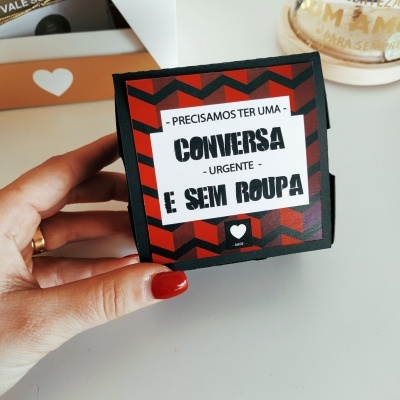 Caixa pequena vermelha e preta com texto em português segurada por mão com unha vermelha