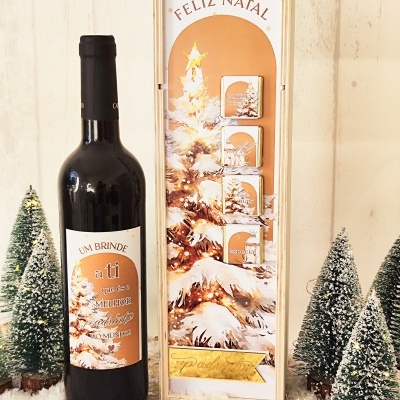Garrafa de vinho tinto e caixa decorativa de Natal com pinheiro nevado e texto FELIZ NATAL, rodeadas por mini árvores de Natal com neve artificial.