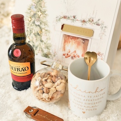 Conjunto natalício com licor, bola de frutos secos, caneca com texto e porta-chaves sobre superfície branca e cartão de Feliz Natal ao fundo