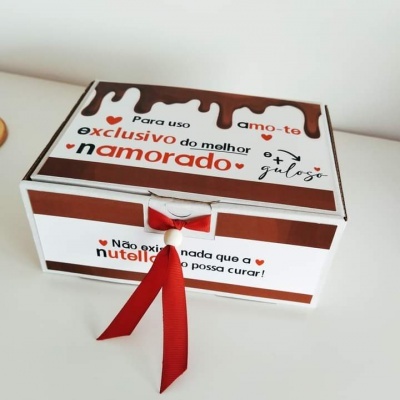 Caixa branca com detalhes marrons e texto em português, com um laço vermelho na frente.