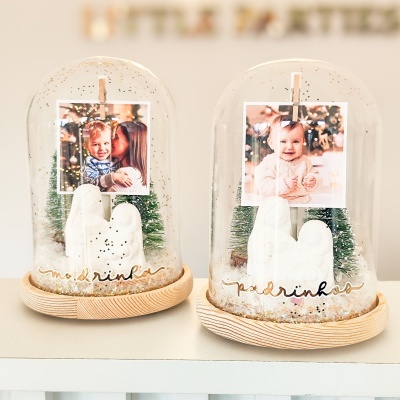 Peças decorativas em cúpulas de vidro com tema natalício, base de madeira, árvores verdes, figuras brancas e fotos de bebés.