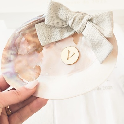 Placa decorativa em concha com laço bege e medalha dourada com letra V