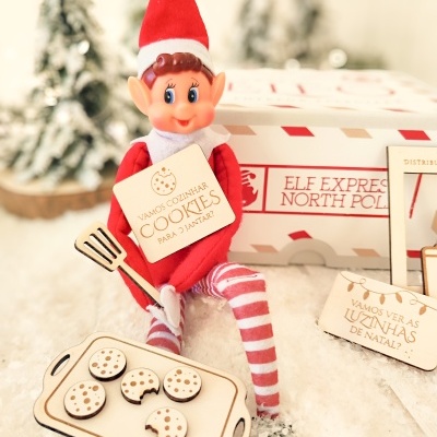 Boneco elfo de Natal com chapéu vermelho e branco sentado na neve com placa e bandeja de bolachas