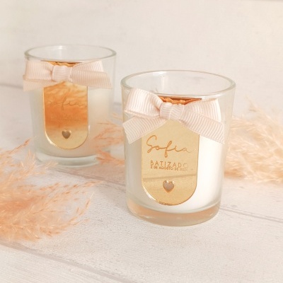 Velas em vasos de vidro com etiqueta dourada e laço rosa claro, sobre superfície branca com planta decorativa.