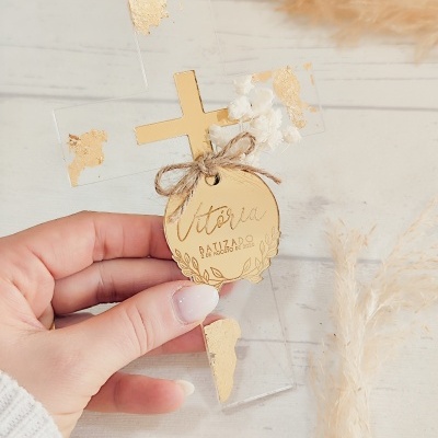 Cruz decorativa transparente com detalhes dourados e etiqueta com texto para batizado.