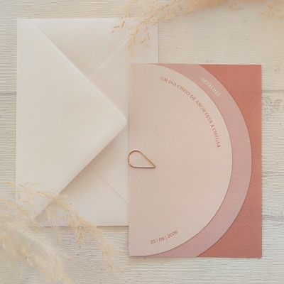 Convite rosa com envelope branco e ramos secos sobre mesa de madeira clara