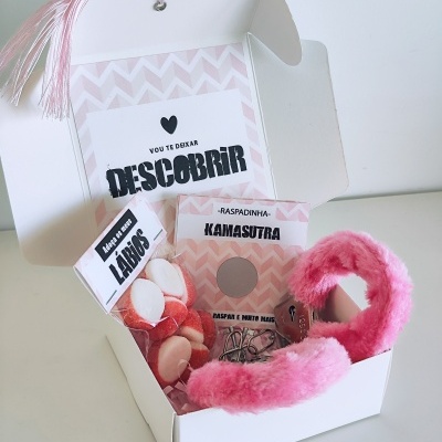 Caixa branca aberta com venda rosa felpuda, confeitos em forma de lábios e cartões com frases em português.