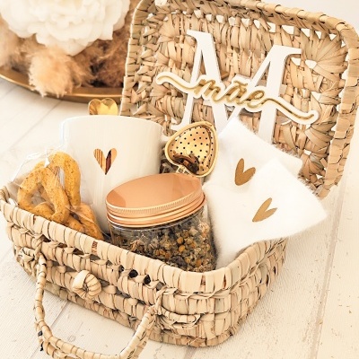 Cesto de palha com caneca branca, frasco com tampa cobre, saquinhos brancos, colher em forma de coração dourado e letra decorativa M com palavra irisé
