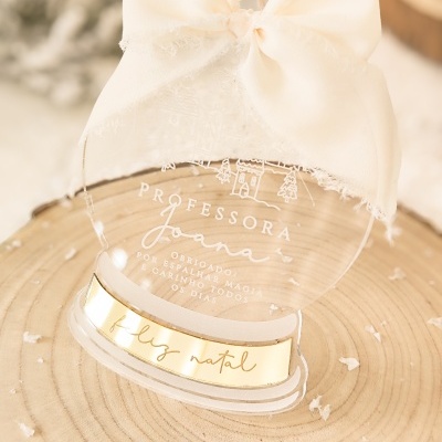 Objeto decorativo transparente redondo com laço creme e texto gravado para professora, sobre tronco de árvore