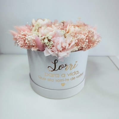 Arranjo floral em caixa branca redonda com flores cor-de-rosa e brancas e texto dourado.